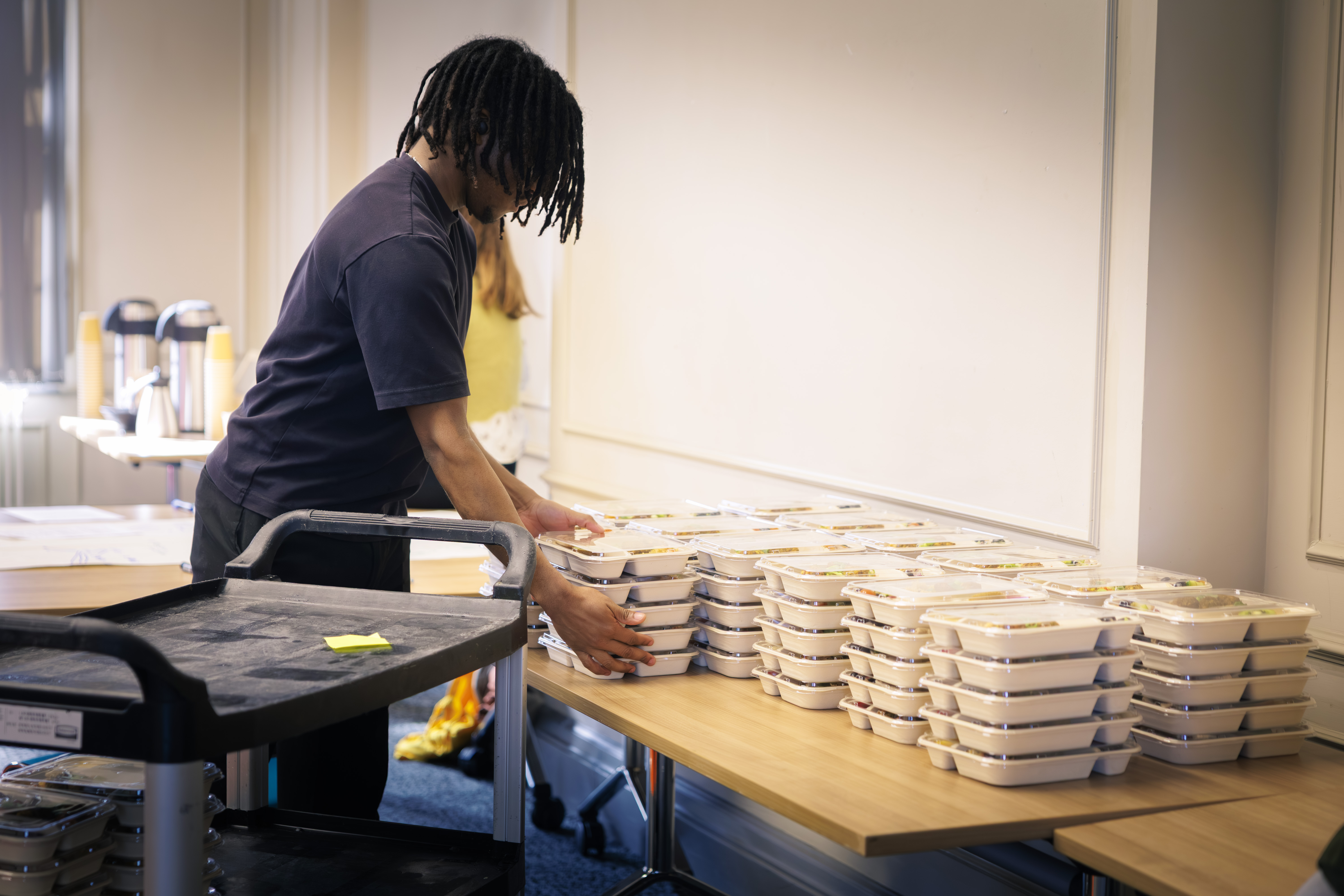 A worker delivers trays of food to a table at the back of a meeting room