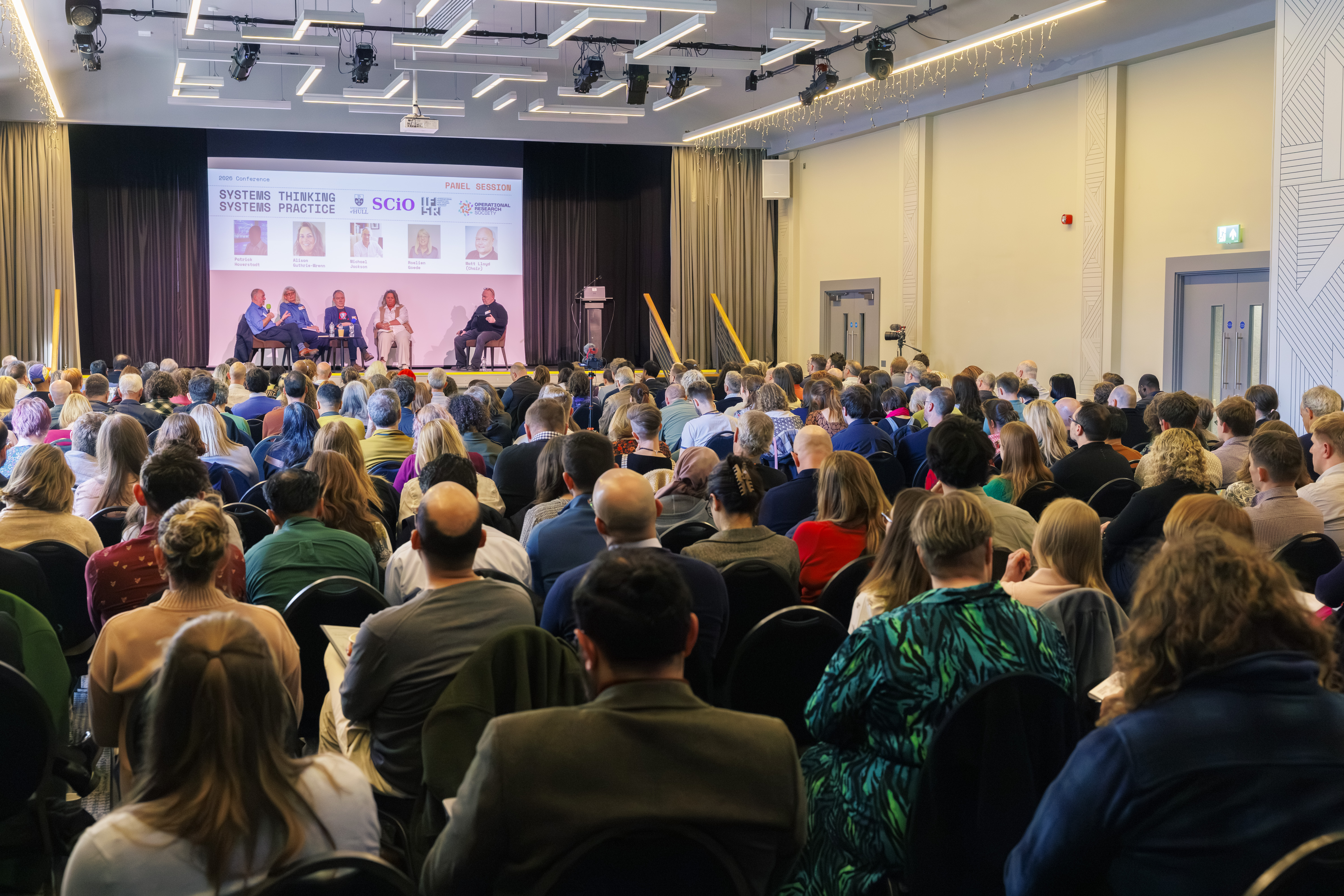 A large group of people watch a panel discussion in a bright, modern room