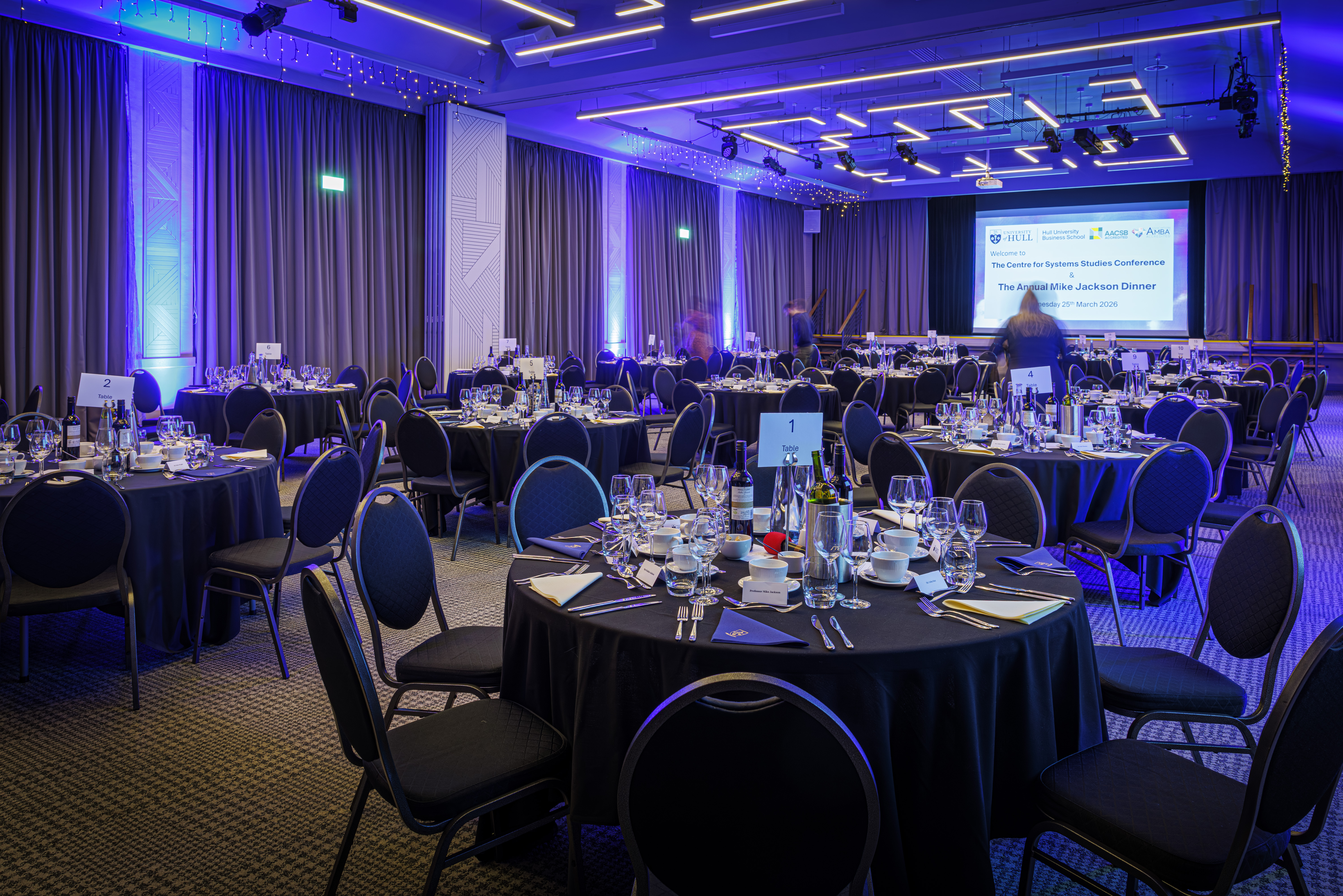A large room with mood lighting, with circular tables set up with formal dinnerware