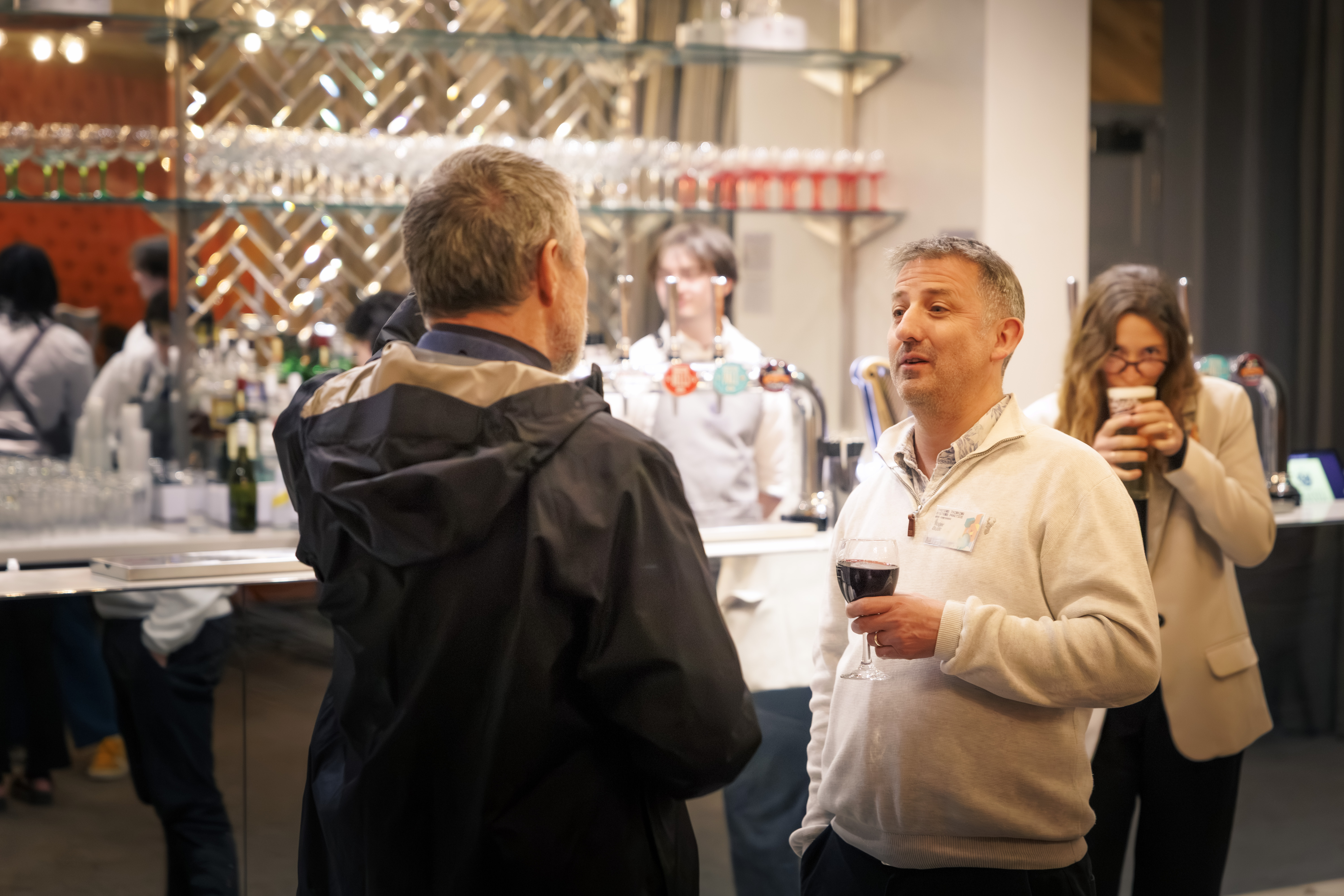 Two people stand talking with a drink in a bar