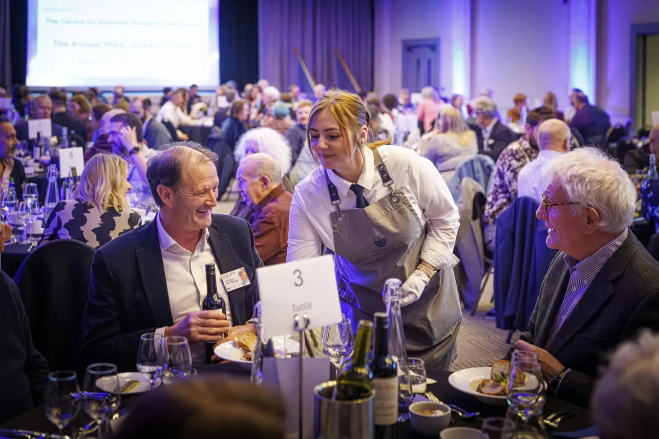A smiling waitress carefully places a plate of food in front of a laughing man at a conference meal