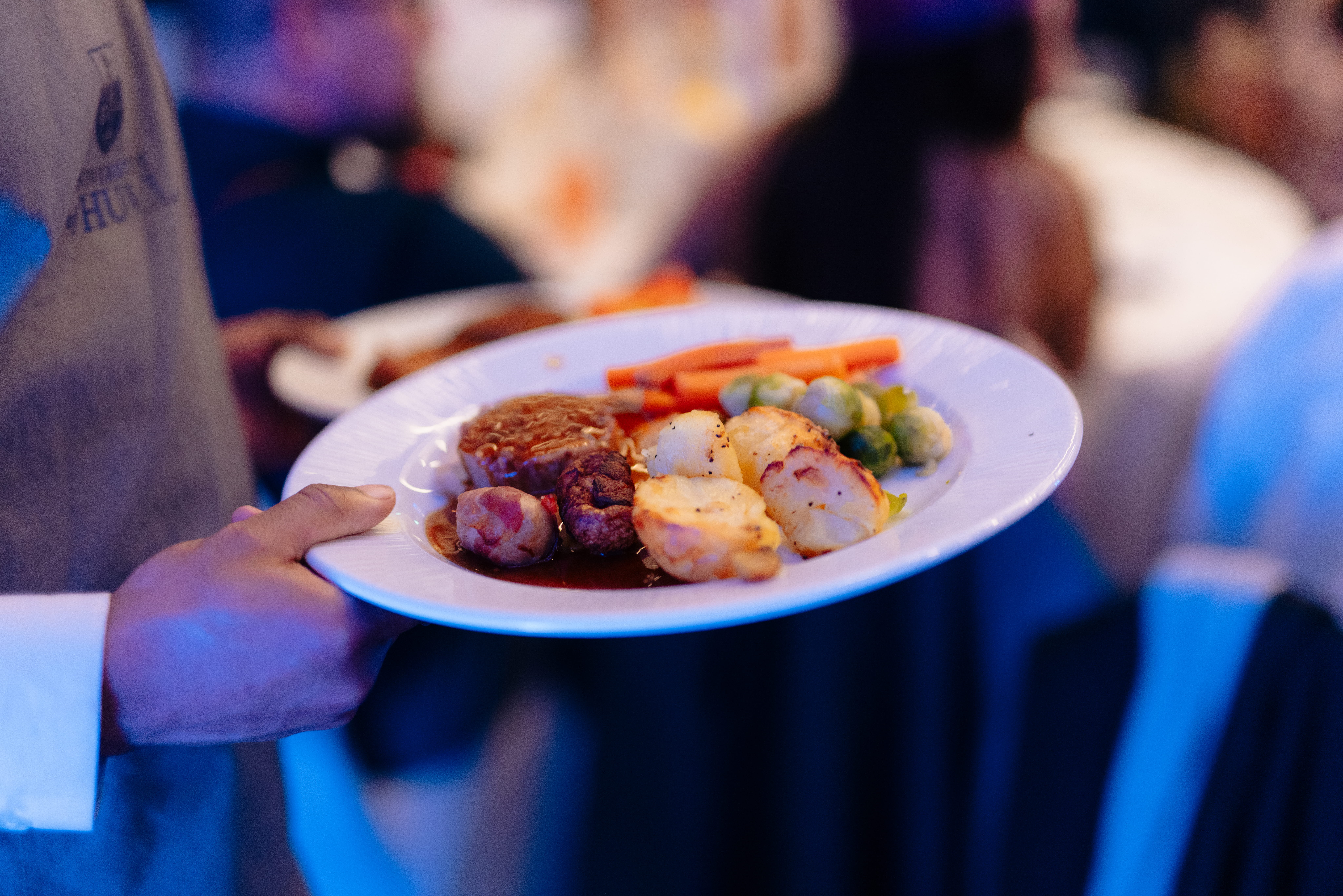 A waiter holding a white plate with beef, roast potatoes, carrots, sprouts and gravy on.