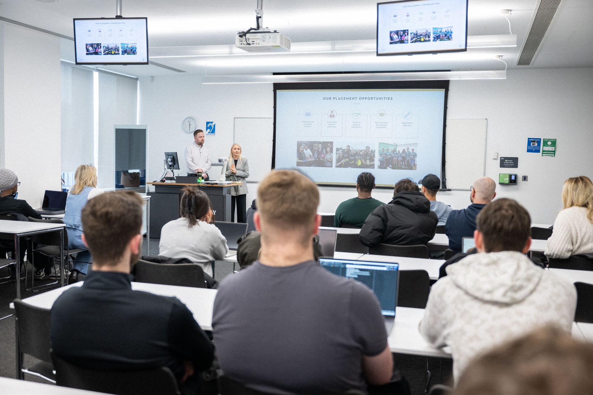 A pair of presenters give a talk in a lecture room with multiple screens showing a presentation