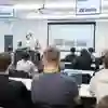 A pair of presenters give a talk in a lecture room with multiple screens showing a presentation
